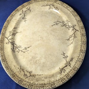 Könnte beinhalten: Eine ovale Keramikplatte mit einer cremefarbenen, gesprenkelten Oberfläche. Der Rand ist mit einem dunkelbraunen geometrischen Muster verziert. Die Mitte und die Ränder zeigen dunkelbraune Illustrationen blühender Zweige.