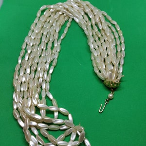 Op de afbeelding: Een meerstrengs parelketting met een gouden sluiting en een enkele parel hanger. De ketting is gemaakt van ovale, nepparels en ligt op een groene ondergrond.
