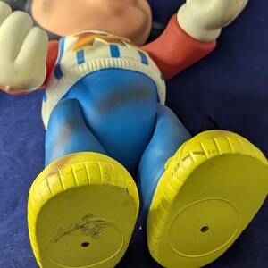 May include: A vintage Mickey Mouse figurine, lying on its back. The figure has a red hat, black ears, and a white and blue sweater. The pants are blue, and the shoes are yellow.