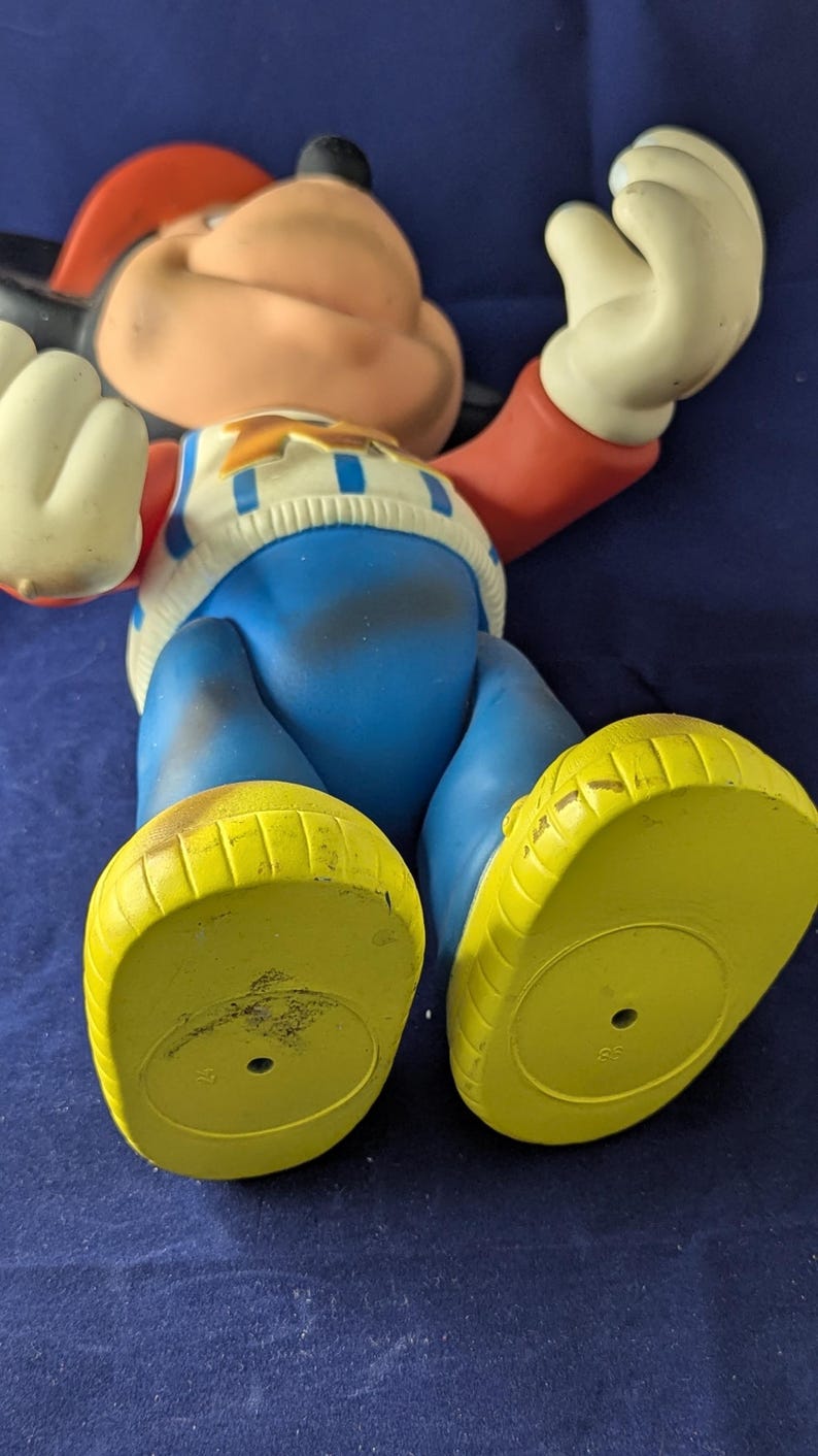 May include: A vintage Mickey Mouse figurine, lying on its back. The figure has a red hat, black ears, and a white and blue sweater. The pants are blue, and the shoes are yellow.
