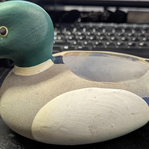 Puede incluir: Un cenicero con forma de pato pintado a mano. El pato tiene la cabeza verde, el pico beige y el cuerpo beige y blanco con detalles azules. El cenicero está hecho de un material sólido y tiene un acabado liso. El pato está sobre una superficie oscura.