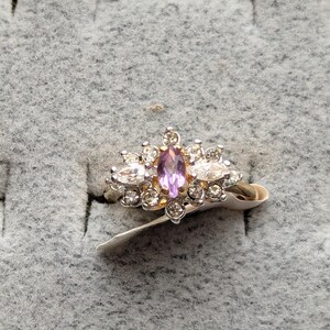 May include: A silver ring with a central oval purple gemstone, surrounded by smaller clear stones. The ring is set against a gray, textured background.
