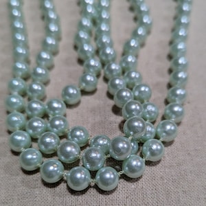Puede incluir: Una hebra de cuentas de imitación de perlas de color verde claro. Las cuentas están enhebradas en un hilo transparente y están dispuestas en un patrón circular.
