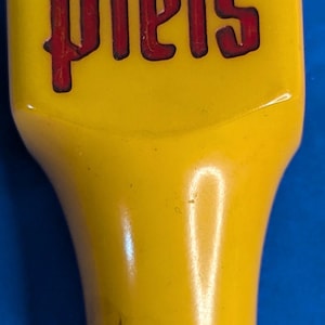 May include: A yellow beer tap handle with the word "Piels" in red lettering. The handle has a rounded top and a curved, elongated body. The background is a solid blue.