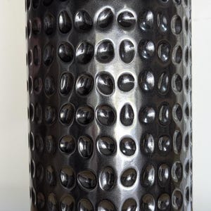 May include: A tall, cylindrical black vase with a hammered metal texture. The surface is covered in evenly spaced, rounded indentations. The vase has a glossy finish and is suitable for home decor.