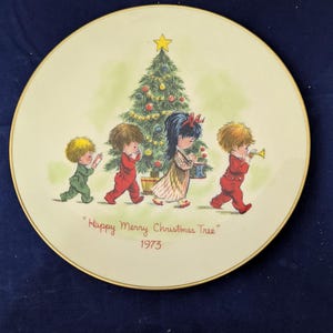 Op de afbeelding: Een decoratieve plaat met een kerstthema. De plaat toont een geschilderde scène van kinderen rond een versierde kerstboom. De tekst "Happy Merry Christmas Tree" en het jaar 1973 staan op de plaat. De plaat heeft een gouden rand.