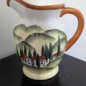 May include: A ceramic jug with a painted landscape scene. The jug has a white base with a brown handle and rim. The landscape depicts rolling green hills, trees, and small houses with red roofs. The jug is a decorative item.