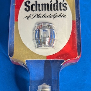 May include: A clear plastic beer tap handle with a red, white and gold design. The handle features the text "Schmidt's of Philadelphia" and a silver metal barrel.