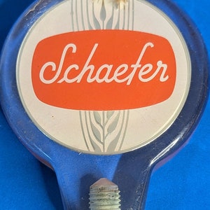 May include: A clear plastic beer tap handle with a blue base and a white circle with a red oval containing the word "Schaefer" in white script. The circle also has a gray wheat design.