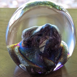 Può includere: Un fermacarte in vetro trasparente con un vortice iridescente scuro all'interno. Il vortice è di colore viola, blu e oro.