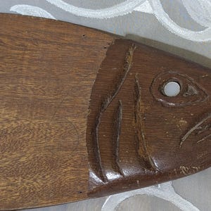 Puede incluir: Una tabla de cortar de madera con forma de pez y acabado marrón oscuro. El pez tiene detalles tallados, incluyendo un ojo, branquias y una boca. La tabla está diseñada para la preparación y el servicio de alimentos.