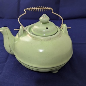 May include: A light green ceramic teapot with a rounded body, a matching lid featuring a small knob, and a curved spout. The teapot includes a metal wire handle with a coiled spring grip attached to two side lugs, and a subtle crackle glaze finish.