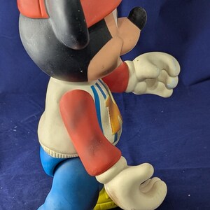 May include: A vintage Mickey Mouse figurine, wearing a red firefighter's hat with a yellow "M". The figure has a white jacket with blue and red details, blue trousers, and yellow shoes. A collectible item.