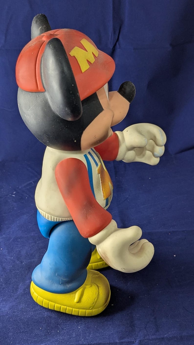 May include: A vintage Mickey Mouse figurine, wearing a red firefighter's hat with a yellow "M". The figure has a white jacket with blue and red details, blue trousers, and yellow shoes. A collectible item.