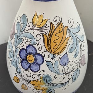 Puede incluir: Jarrón de cerámica blanca con diseño floral pintado a mano. El jarrón presenta un borde azul y un patrón detallado de flores amarillas, azules y moradas con hojas verdes y espirales negras. El jarrón tiene forma redondeada.