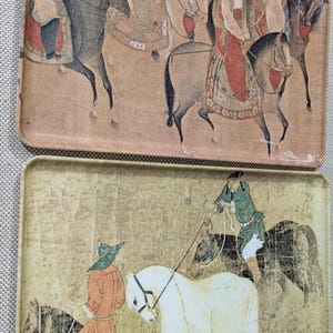 May include: Two rectangular serving trays featuring traditional East Asian art. The top tray depicts figures on horseback in muted earth tones. The bottom tray features a gold-toned background with figures riding horses, including a prominent white horse.
