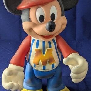May include: A vintage Mickey Mouse figurine. The figure wears a red cap with a yellow "M", a red and white striped shirt with a yellow "M", blue trousers, and yellow shoes. The figure has black ears and white gloves.