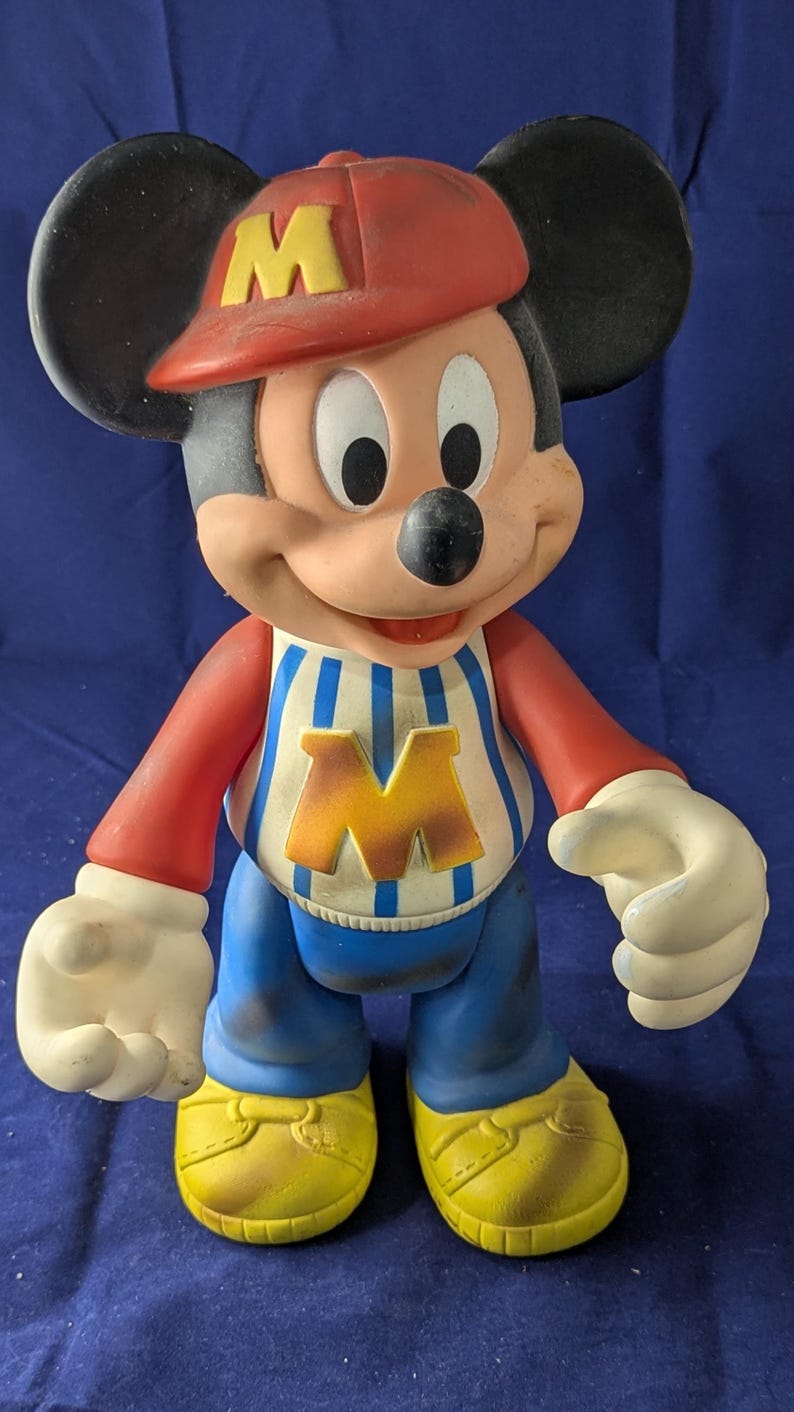 May include: A vintage Mickey Mouse figurine. The figure wears a red cap with a yellow "M", a red and white striped shirt with a yellow "M", blue trousers, and yellow shoes. The figure has black ears and white gloves.
