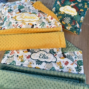 Peut inclure: Une couverture bébé verte et jaune avec un imprimé d'animaux de la jungle. La couverture a deux couches, l'une en polaire jaune et l'autre en polaire verte. La couverture est personnalisée avec les noms "Louane" et "Elio".