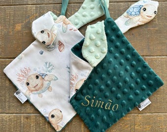 May include: Two baby security blankets. One is white with a sea turtle and coral pattern, the other is dark green with a dimpled texture. Both have a green loop and a turtle-shaped attachment. The green blanket has the name "Simão" embroidered in gold.