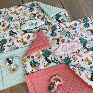 Peut inclure: Deux couvertures pour b&eacute;b&eacute; personnalis&eacute;es avec un imprim&eacute; d'animaux de la jungle. Une couverture est bleu clair avec le nom "Joy" brod&eacute; dessus. L'autre couverture est rose avec le nom "Lison" brod&eacute; dessus.