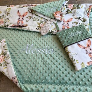 Peut inclure: Une couverture minky vert sauge douce et des accessoires assortis avec un panneau en tissu blanc orné d'un motif répétitif de lapins bruns, de feuilles vertes et de fleurs roses. Le prénom Alessio est brodé en écriture blanche sur le tissu.