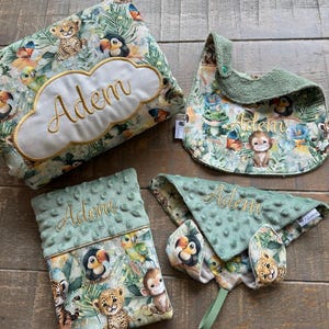 Peut inclure: Un ensemble d'articles pour bébé sur le thème de la jungle. L'ensemble comprend un oreiller vert et marron avec le nom "Adem" brodé dessus, une bavette verte avec le nom "Adem" brodé dessus, une  couverture anti-régurgitation verte et marron avec le nom "Adem" brodé dessus, et une couverture pour bébé verte et marron avec le nom "Adem" brodé dessus.