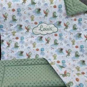 May include: A baby blanket featuring a whimsical dragon pattern in shades of green, blue, and orange on a white background. The blanket includes a green, textured border and a cloud-shaped patch embroidered with the name "Louis".