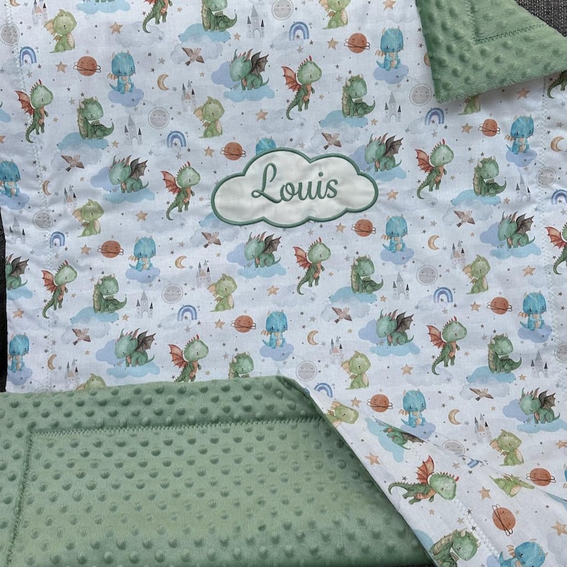 May include: A baby blanket featuring a whimsical dragon pattern in shades of green, blue, and orange on a white background. The blanket includes a green, textured border and a cloud-shaped patch embroidered with the name "Louis".