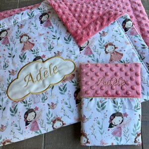 May include: A baby blanket and matching pillow with a fairy theme. The blanket and pillow feature a white background with a pattern of fairies, flowers, and butterflies. The blanket has a pink, textured top, and the pillow has the name "Adele" embroidered on it.