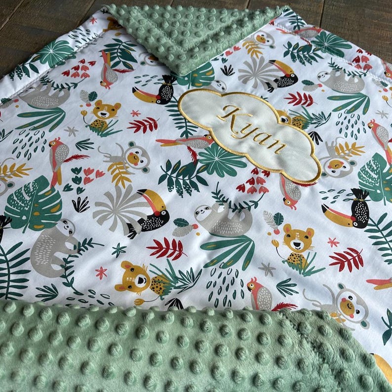 May include: A baby blanket with a white background featuring a jungle animal print, including toucans, sloths, and monkeys. The blanket has a green minky dot border and a cloud-shaped patch embroidered with the name "Ryan" in gold thread.
