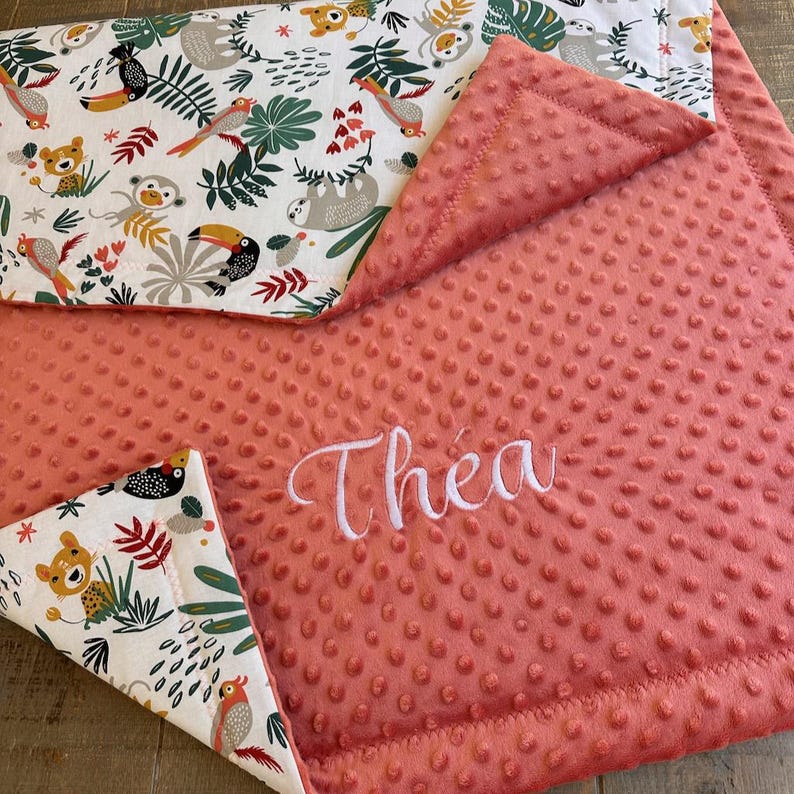 May include: Coral baby blanket with a white embroidered name, "Thea." The blanket features a patterned border with toucans, sloths, monkeys, and other jungle animals. The blanket is made of soft, textured material.