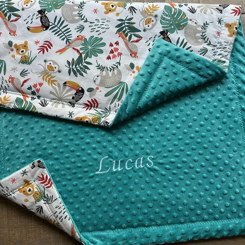 May include: A teal baby blanket with a white and colorful jungle animal print. The blanket features a soft, textured surface and the name "Lucas" embroidered in white.