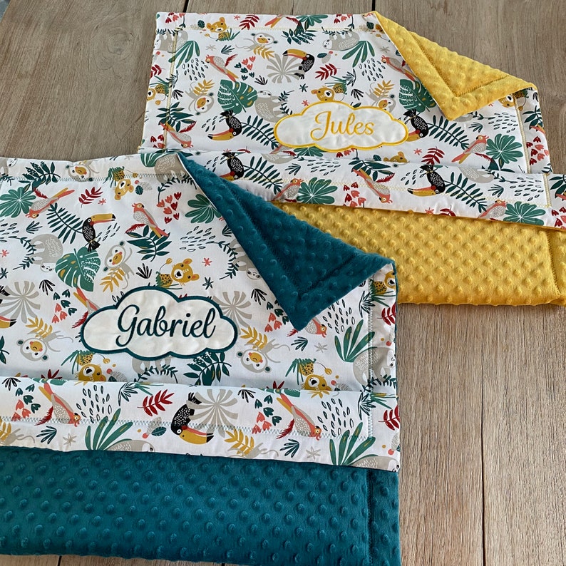 Peut inclure: Deux couvertures pour b&eacute;b&eacute; personnalis&eacute;es avec un imprim&eacute; d'animaux de la jungle. La couverture du haut est jaune avec le nom "Jules" brod&eacute; dessus. La couverture du bas est verte avec le nom "Gabriel" brod&eacute; dessus.