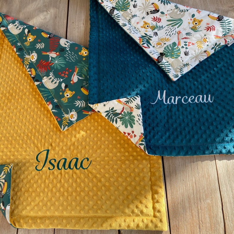May include: Two baby blankets with a jungle theme. One is teal with a textured surface, the other is yellow with a textured surface. Both have a patterned border with toucans, sloths, and tropical leaves. One blanket is embroidered with the name "Isaac", the other with "Marceau".