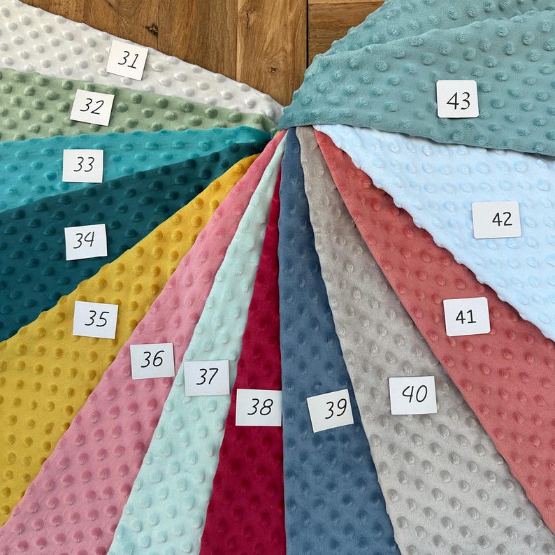 May include: A fan of various colors of dimple dot minky fabric. The colors include white, green, teal, yellow, pink, light blue, red, gray, blue, and coral. Each fabric swatch has a unique number on a white tag.