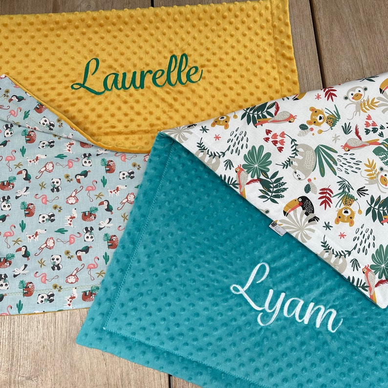 May include: Three baby blankets with different patterns and colors. One is yellow with the name "Laurelle" embroidered in green. Another is teal with the name "Lyam" embroidered in white. The third has a pattern of animals and plants.
