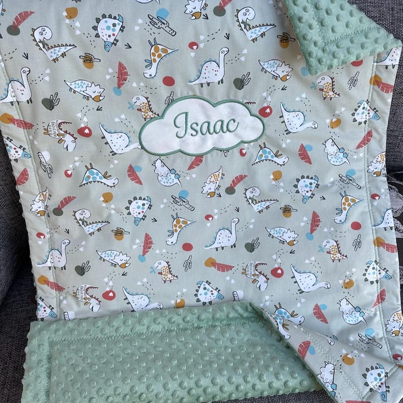 May include: A light green baby blanket with a dinosaur pattern. The blanket features various colorful dinosaur illustrations and the name "Isaac" embroidered in a cloud-shaped patch. The edges are trimmed with a soft, textured green fabric.