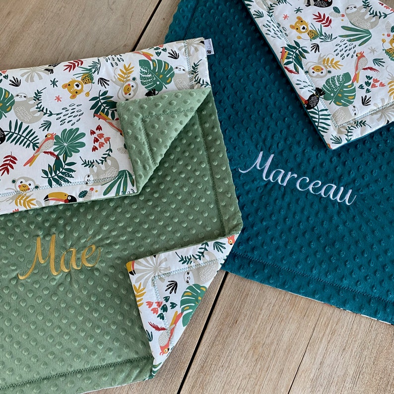 May include: Three baby blankets with jungle animal patterns and personalized names. One blanket is teal, another is green, and the third is white with a jungle print. The names "Mae" and "Marceau" are embroidered.