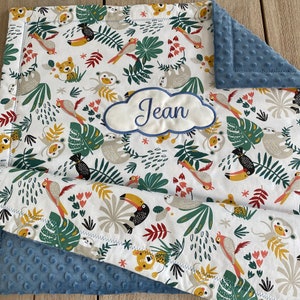 Peut inclure: Une couverture de b&eacute;b&eacute; blanche avec un imprim&eacute; d'animaux de la jungle repr&eacute;sentant des toucans, des paresseux, des singes et des tigres. La couverture est doubl&eacute;e d'un tissu minky bleu et comporte un nuage brod&eacute; personnalis&eacute; avec le nom "Jean".