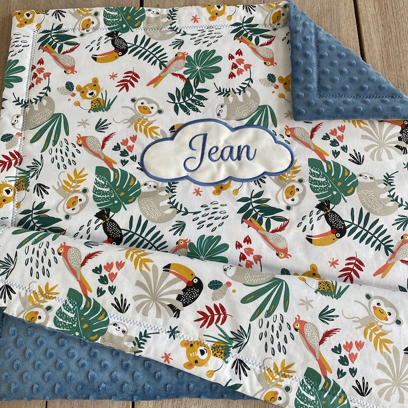 Peut inclure: Une couverture de b&eacute;b&eacute; blanche avec un imprim&eacute; d'animaux de la jungle repr&eacute;sentant des toucans, des paresseux, des singes et des tigres. La couverture est doubl&eacute;e d'un tissu minky bleu et comporte un nuage brod&eacute; personnalis&eacute; avec le nom "Jean".