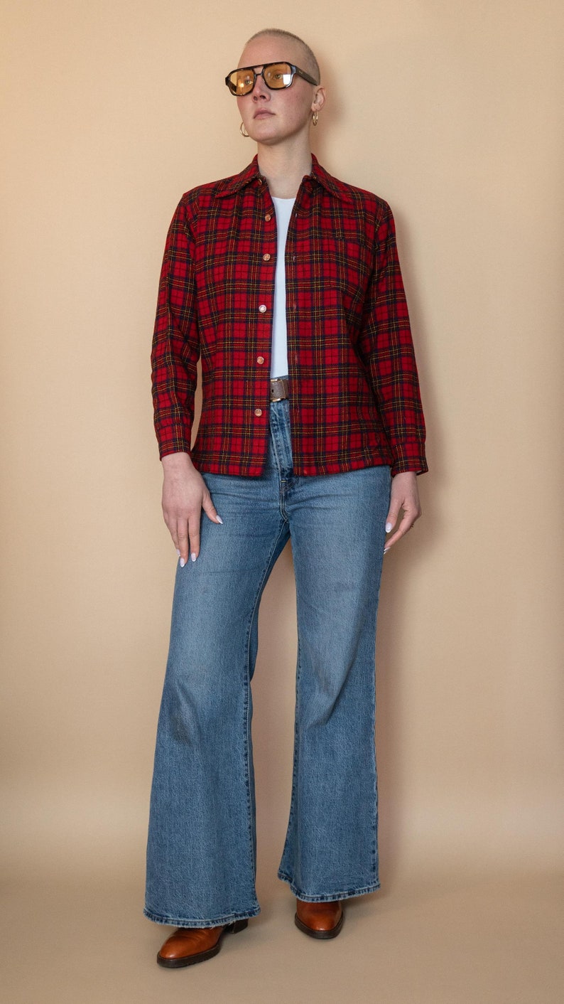 Puede incluir: Una persona lleva una camisa de cuadros rojos y negros con una camiseta blanca debajo, pantalones vaqueros azules y zapatos marrones. Lleva gafas de color amarillo.