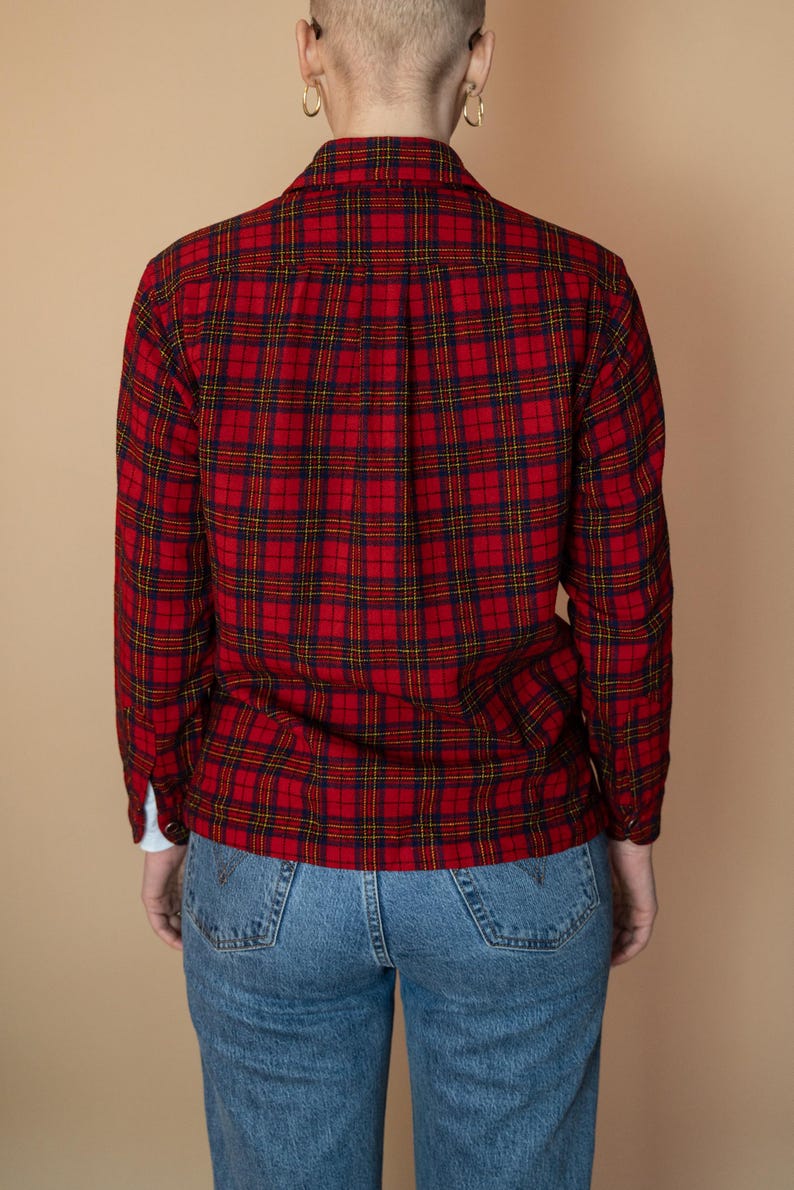 Puede incluir: Una camisa de franela a cuadros rojos y negros con mangas largas y cuello abotonado. La camisa se lleva sobre un par de pantalones vaqueros azules.