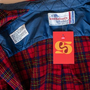 Puede incluir: Camisa de franela a cuadros rojos y azules con forro acolchado azul. La camisa tiene una etiqueta que dice "knockabouts by Pendleton 100% Virgin Wool" y una etiqueta de precio que dice "5 &euro;".