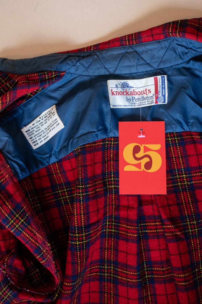 Puede incluir: Camisa de franela a cuadros rojos y azules con forro acolchado azul. La camisa tiene una etiqueta que dice "knockabouts by Pendleton 100% Virgin Wool" y una etiqueta de precio que dice "5 &euro;".