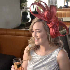 May include: A woman wearing a silver dress and a large, red, fabric flower hat. She is holding a glass of champagne and smiling. The text "Feather Finesse" is visible in the lower right corner of the image.