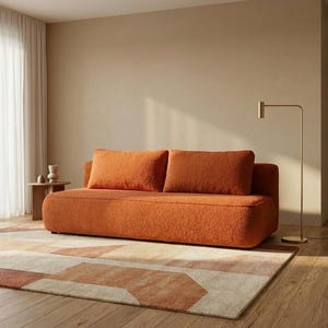 Op de afbeelding: Een oranje bank met twee bijpassende kussens staat op een geometrisch patroon tapijt in een woonkamer. Het tapijt heeft beige, bruine en oranje tinten. Een gouden vloerlamp en een kleine bijzettafel zijn ook zichtbaar.