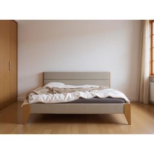 Op de afbeelding: Een modern bedframe met een lichtbeige gestoffeerd hoofdbord en houten poten. Het bed is opgemaakt met wit en tan beddengoed, inclusief een dekbed en kussens. De kamer heeft hardhouten vloeren en een neutrale muurkleur.