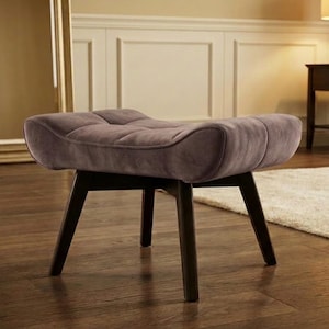 May include: A mauve upholstered ottoman featuring a tufted, curved seat cushion supported by four dark brown wooden legs. The seat has a rectangular shape with rounded edges and a soft, textured fabric finish, providing a stable base.