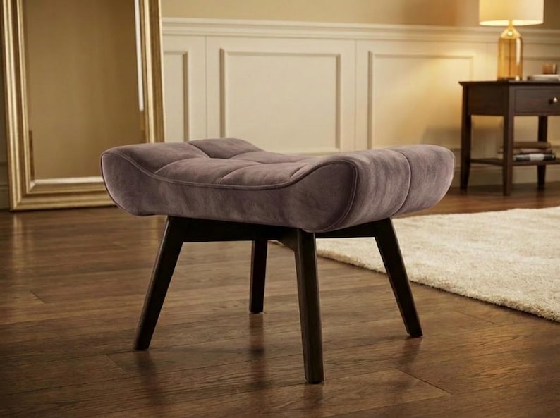 May include: A mauve upholstered ottoman featuring a tufted, curved seat cushion supported by four dark brown wooden legs. The seat has a rectangular shape with rounded edges and a soft, textured fabric finish, providing a stable base.